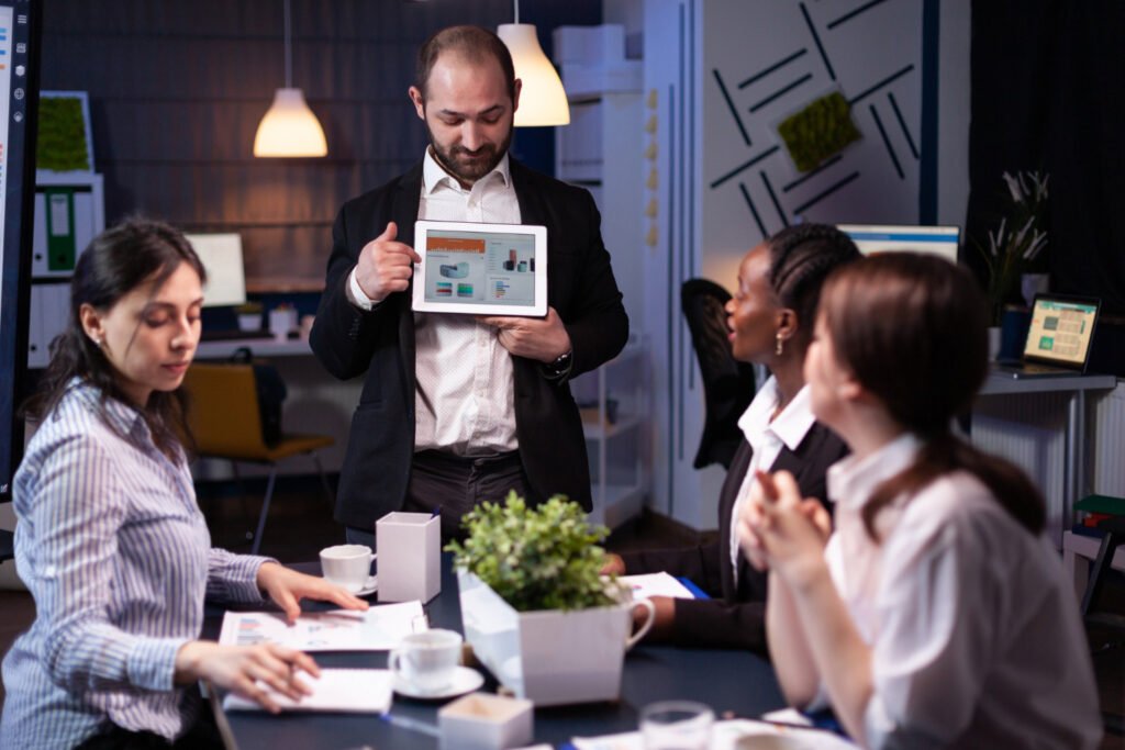 Team leader standing and showcasing presentation metrics and graphics on a tablet screen to seated coworkers during a nighttime office digital marketing Bristol strategy session.